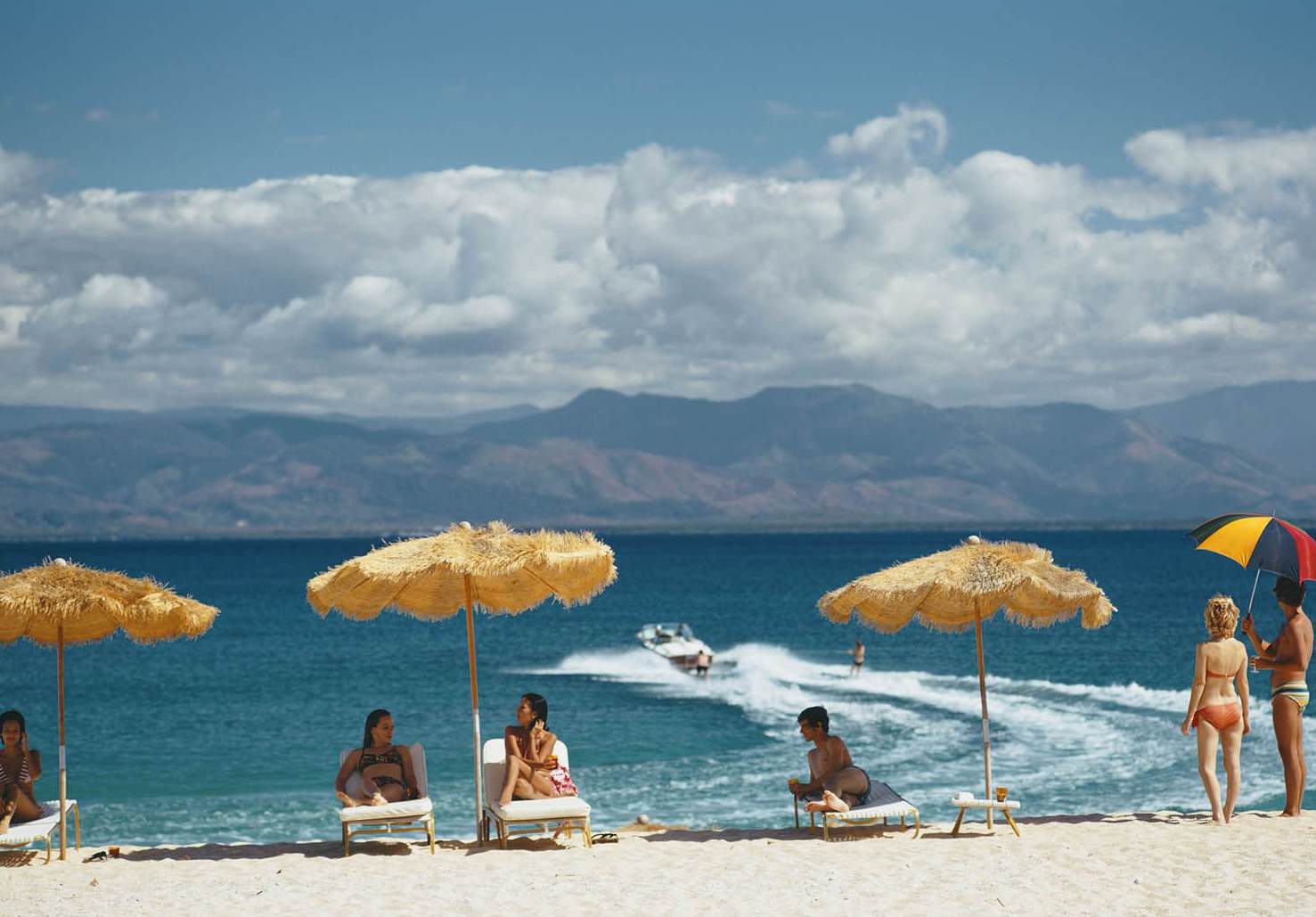 Archival beach scene with guests relaxing beneath thatched umbrellas as a boat passes offshore.