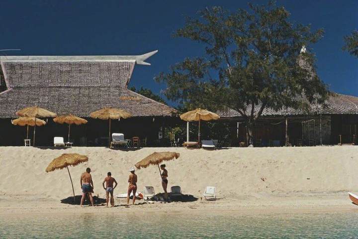 Guests gather beneath thatched umbrellas on the Main Beach, with the beach house behind them.