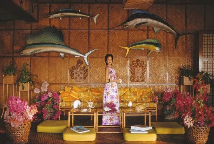 Mary Lou Lovina in a floral dress, surrounded by flowers, bamboo furniture and mounted fish in an island interior.