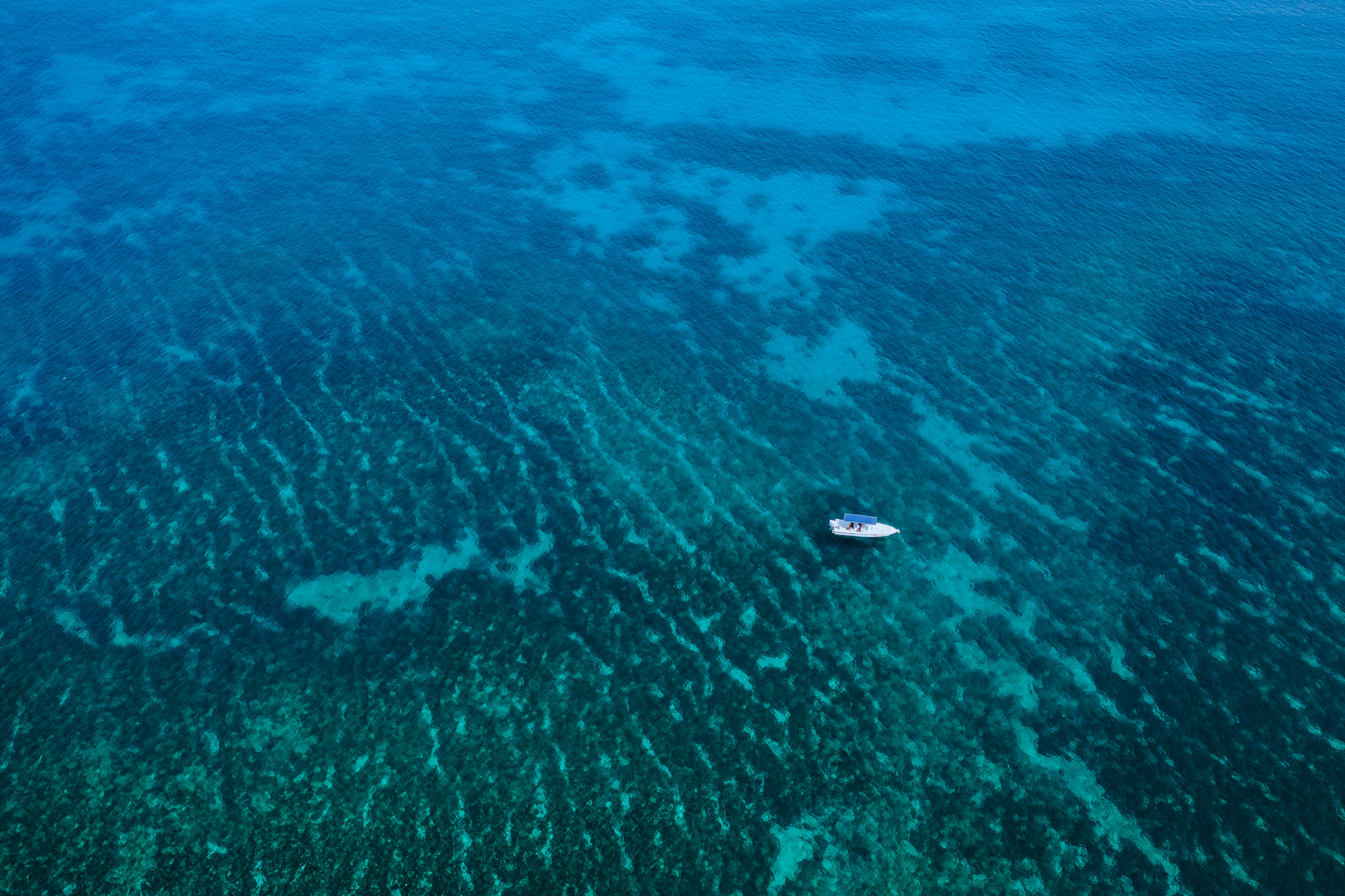A small white boat floats above a patchwork of reef and clear blue water.