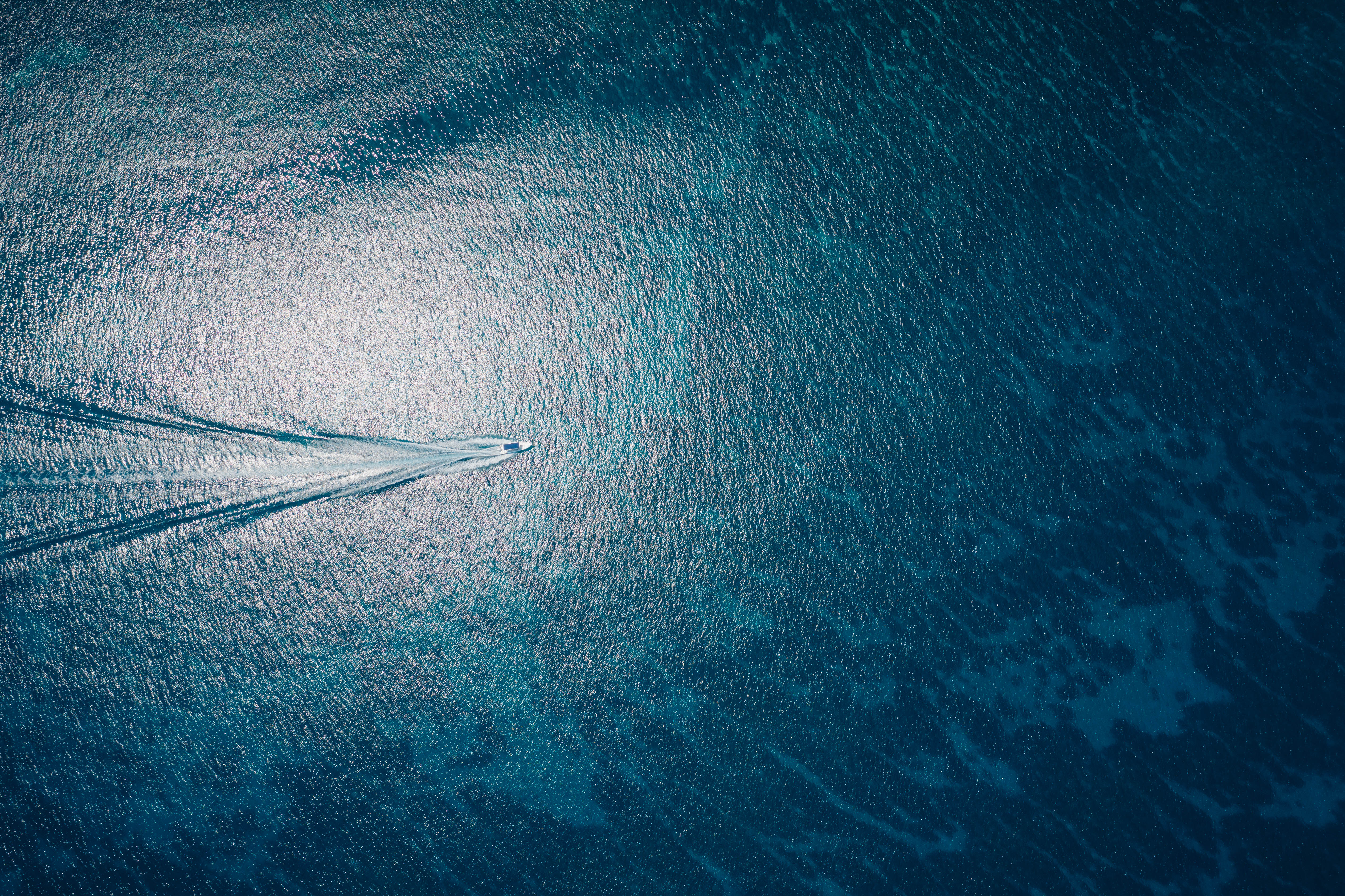 A white boat leaves a long wake across sunlit blue water, seen from above.