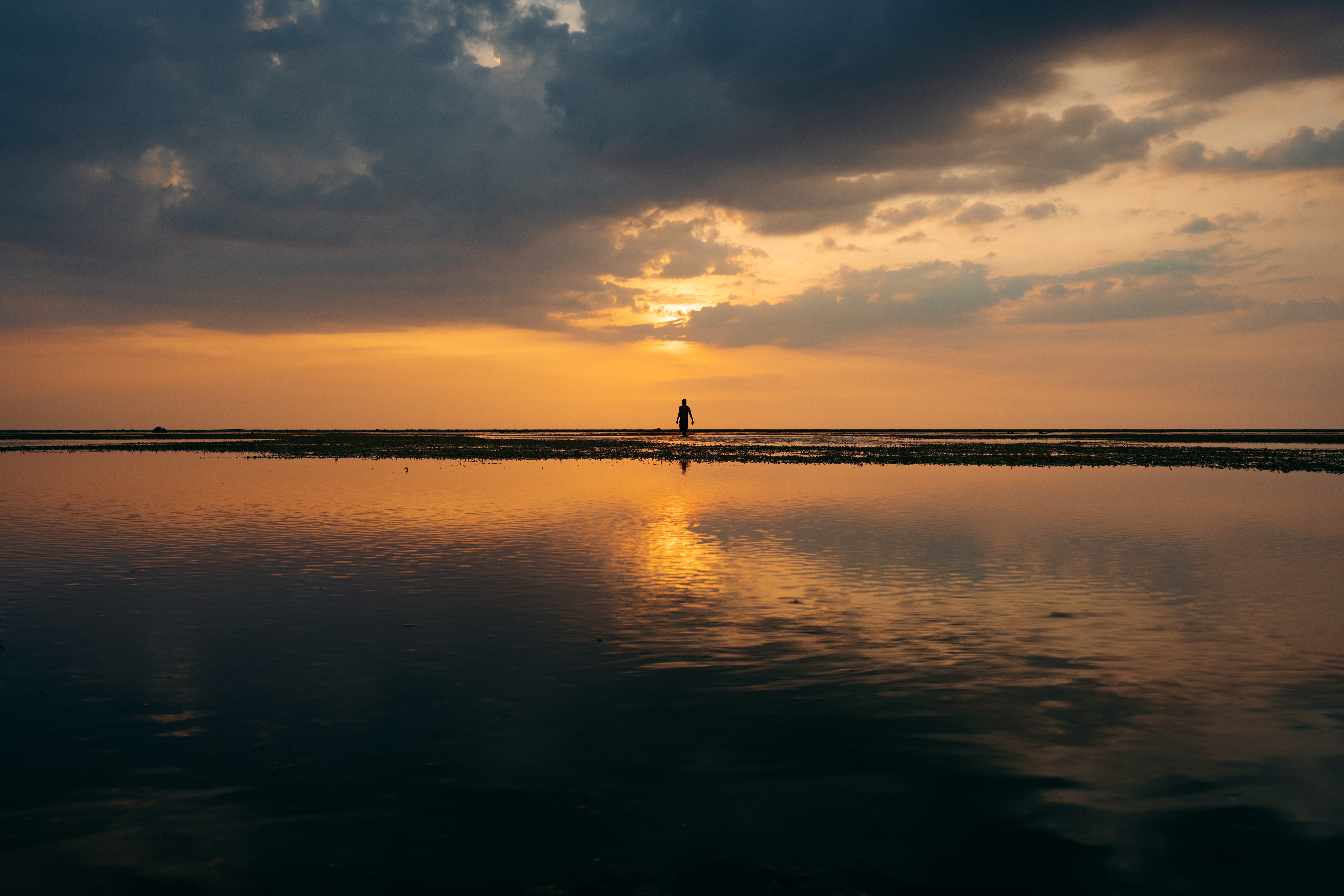 A lone figure on the sandbar beneath a dramatic sunset, with gold light reflected across the still water.