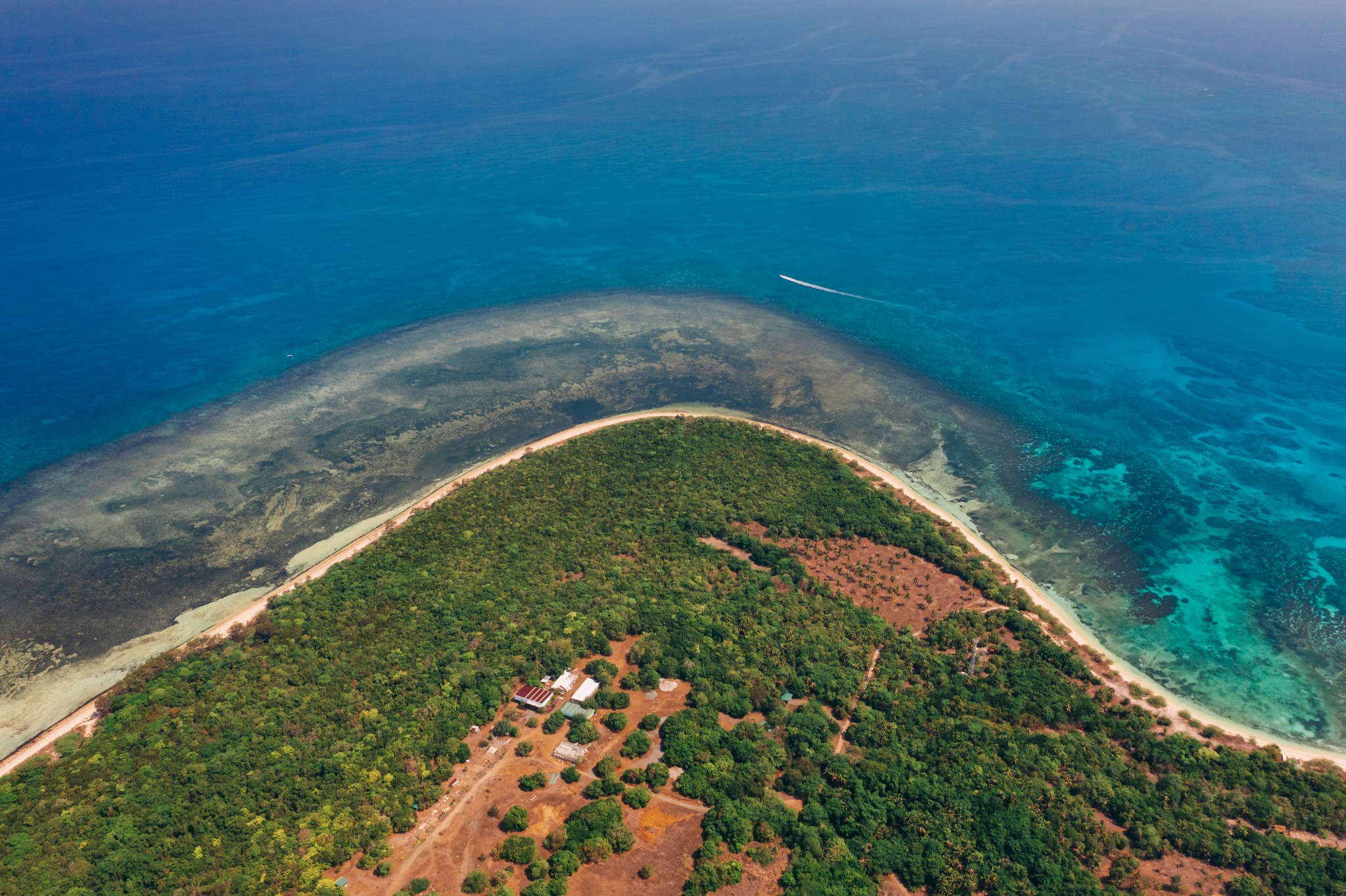 Actual aerial photograph of the curved Hermana Mayor beachfront, with forest, a broad reef and turquoise sea.