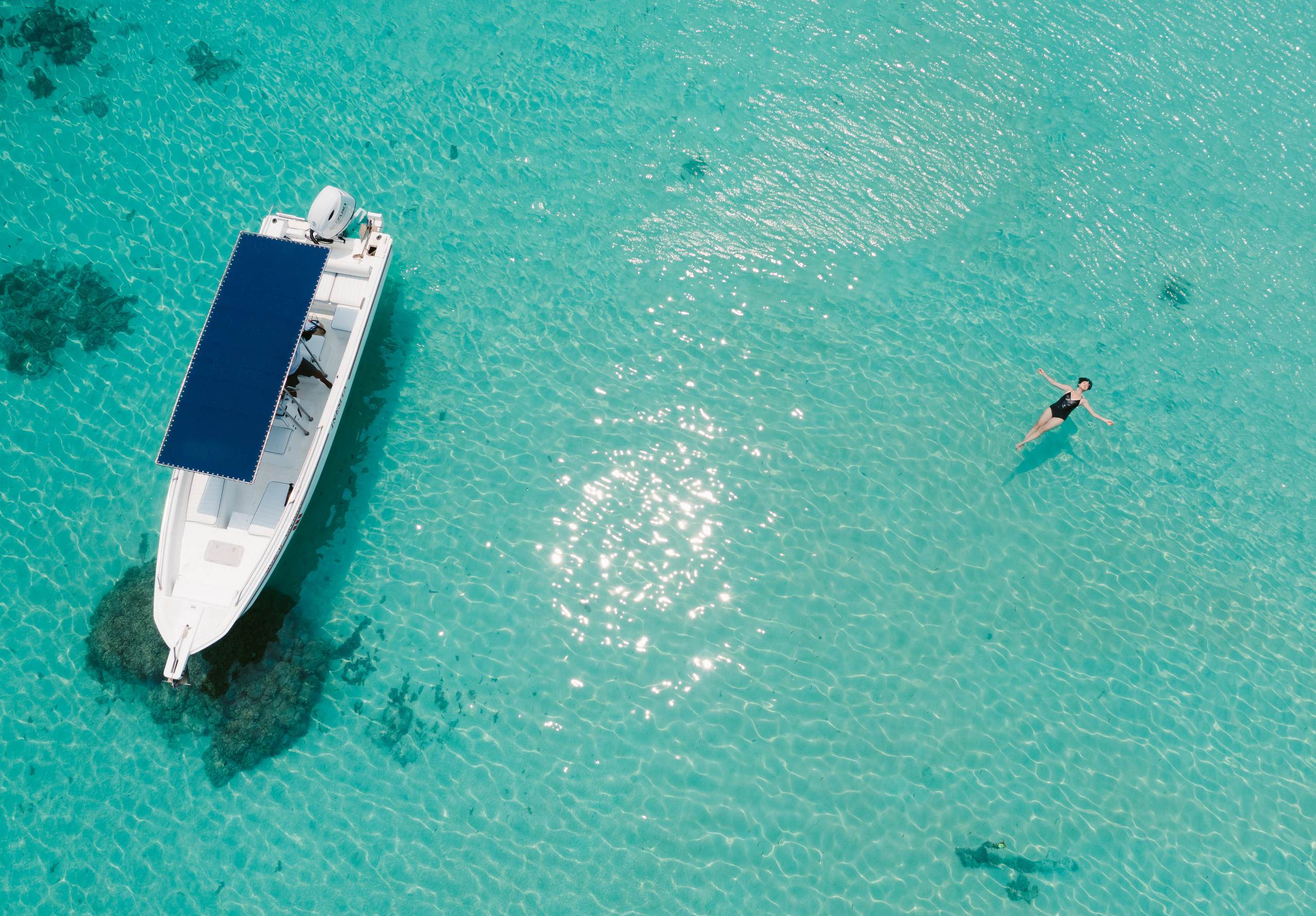 An overhead photograph of a white boat and a swimmer floating in clear turquoise shallows.