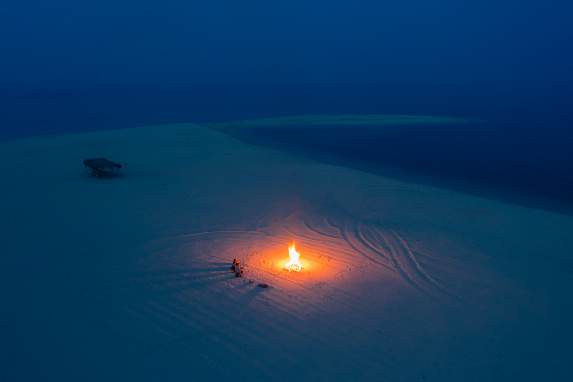 A small bonfire glows on the sandbar against the deep blue sea after dark.