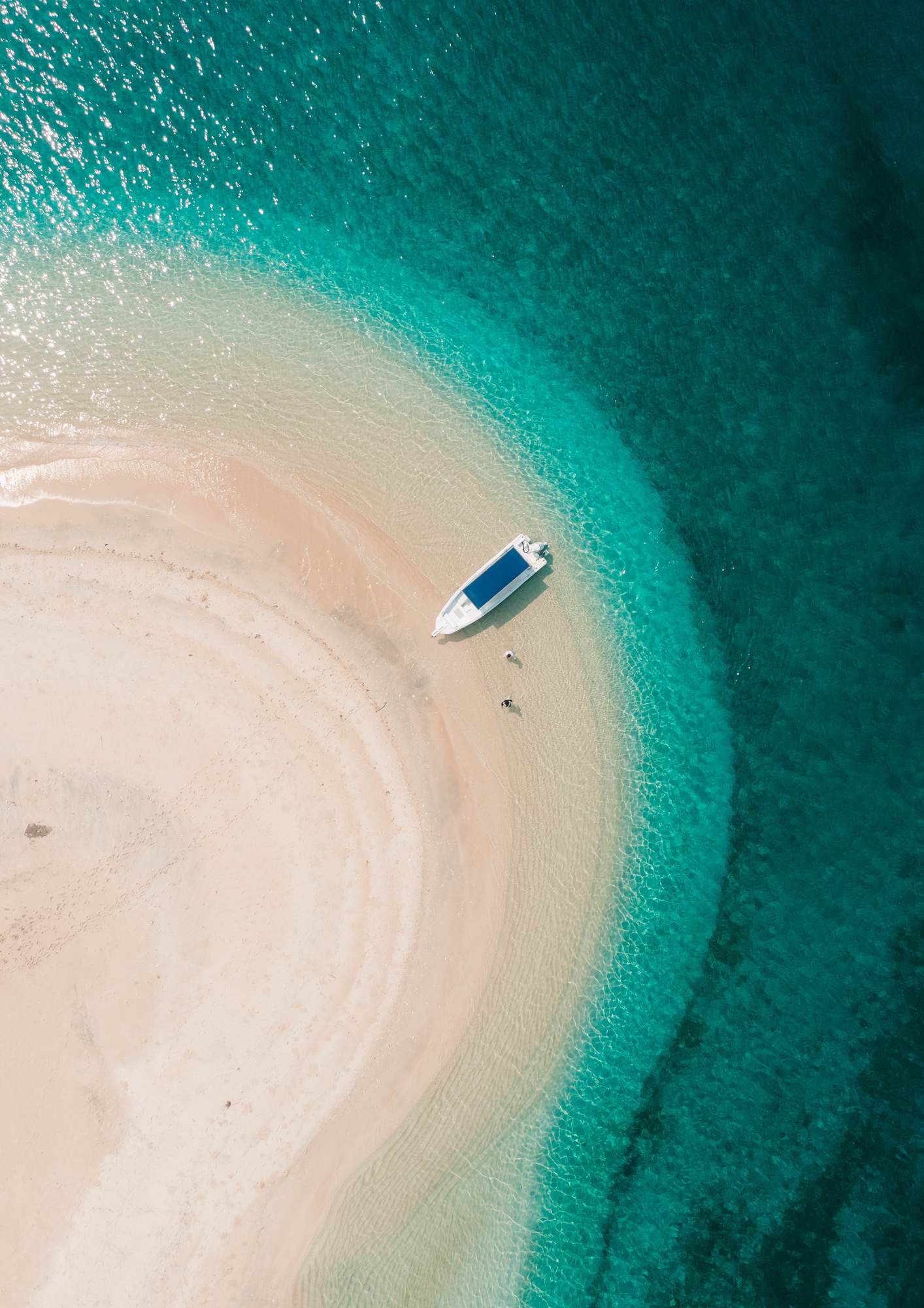 A white boat rests beside the curving sandbar, with two visitors in the shallow turquoise water.