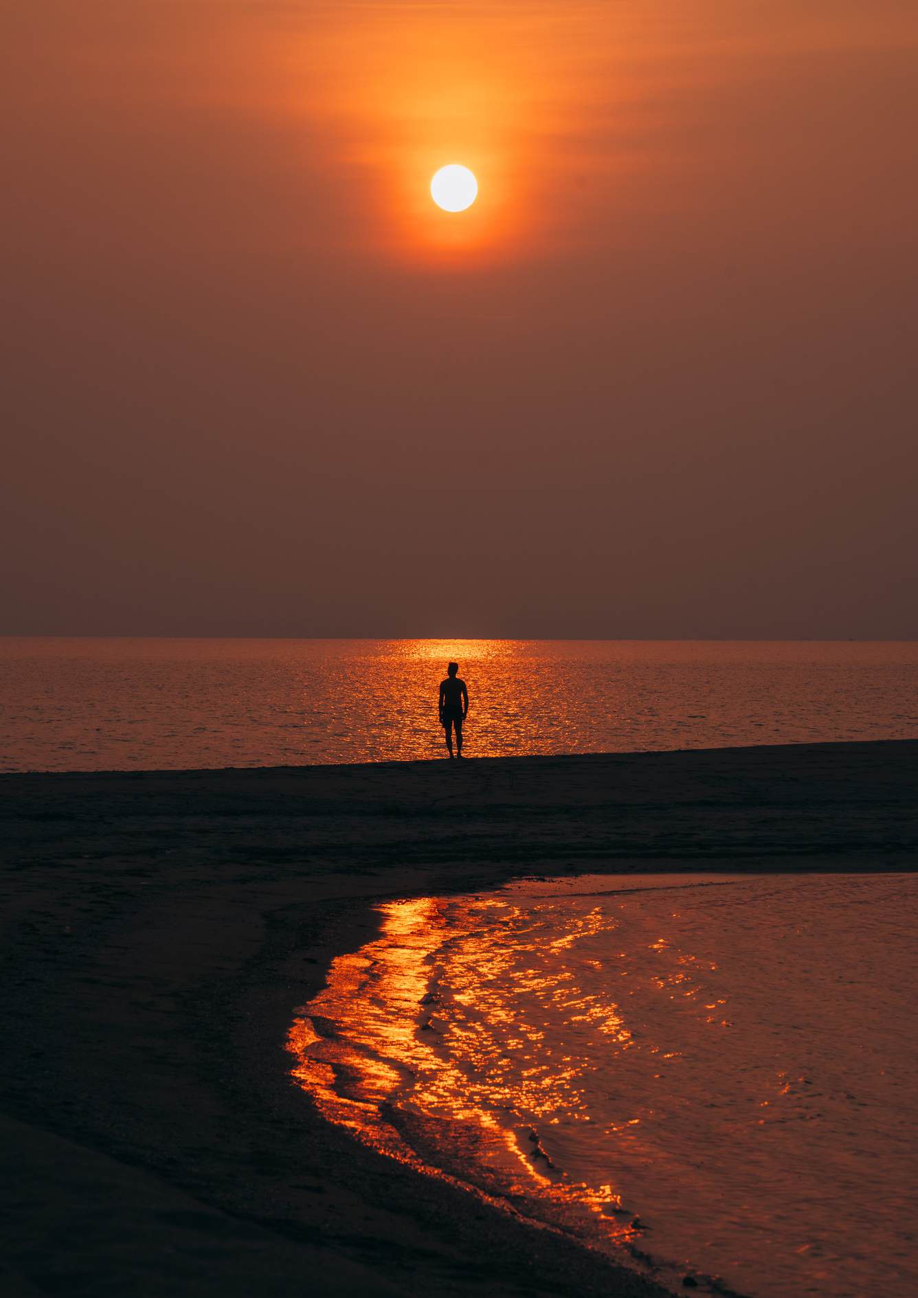 A lone figure stands on the sandbar beneath an orange sunset, reflected in the sea and shallow water.